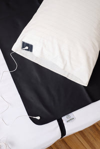 Earthle Erdungskissenbezug auf einem Bett neben der schwarzen Erdungsschlafmatte, mit dem Erdungskabel daran